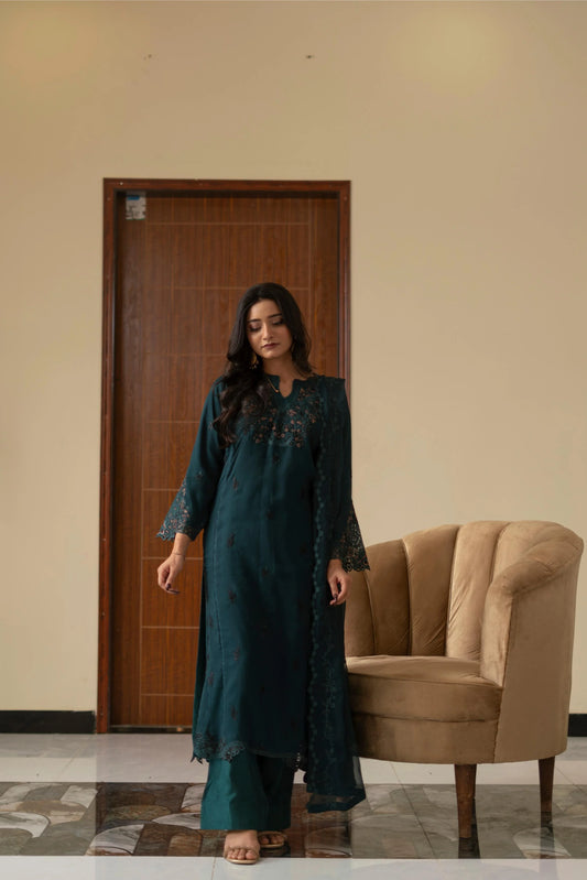 Woman in a teal outfit standing in a room with a beige wall and brown chair.