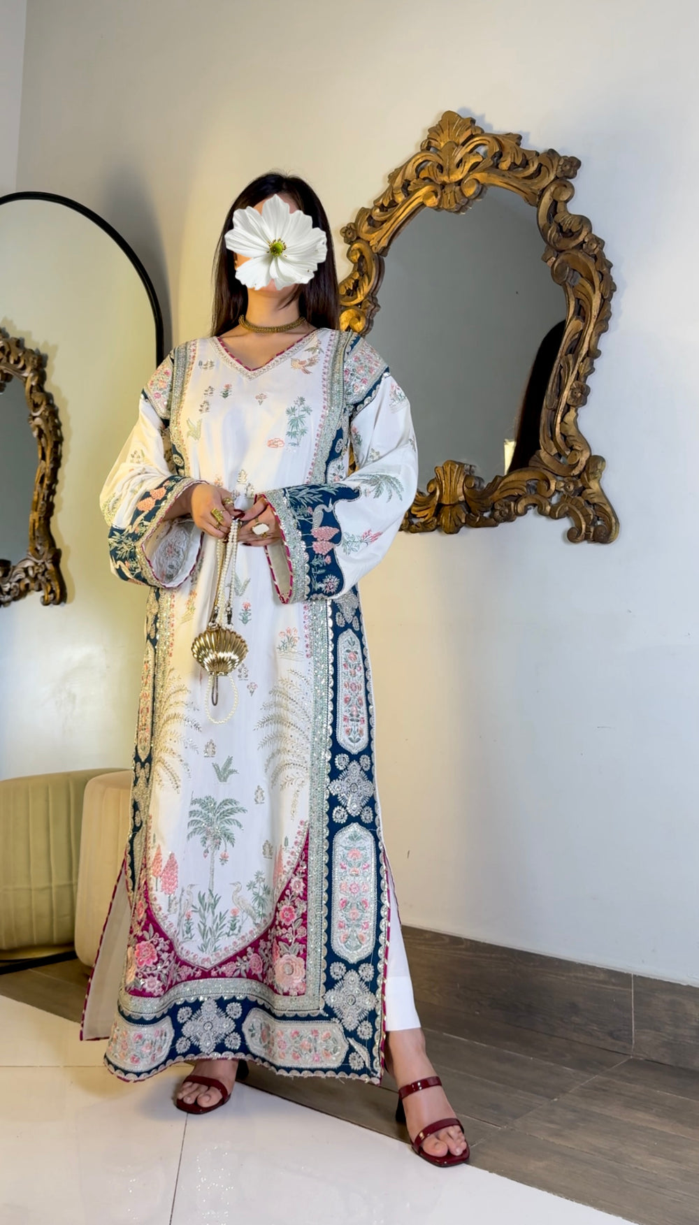 A woman wearing a long white embroidered kurta with blue and pink border patterns, standing indoors in front of ornate mirrors.