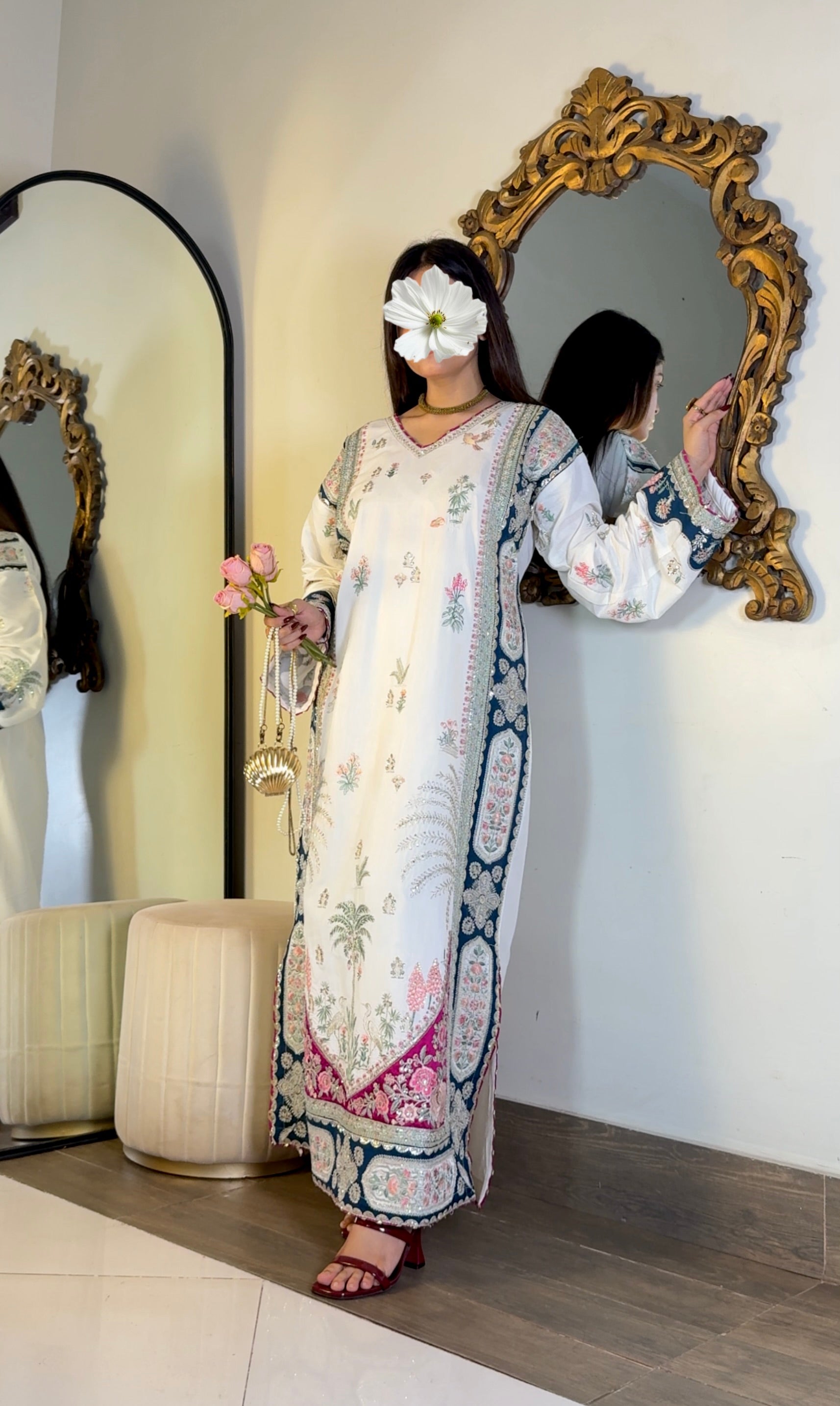 A woman models a long white embroidered dress with blue and pink borders, standing in front of a large ornate gold mirror in an indoor setting.
