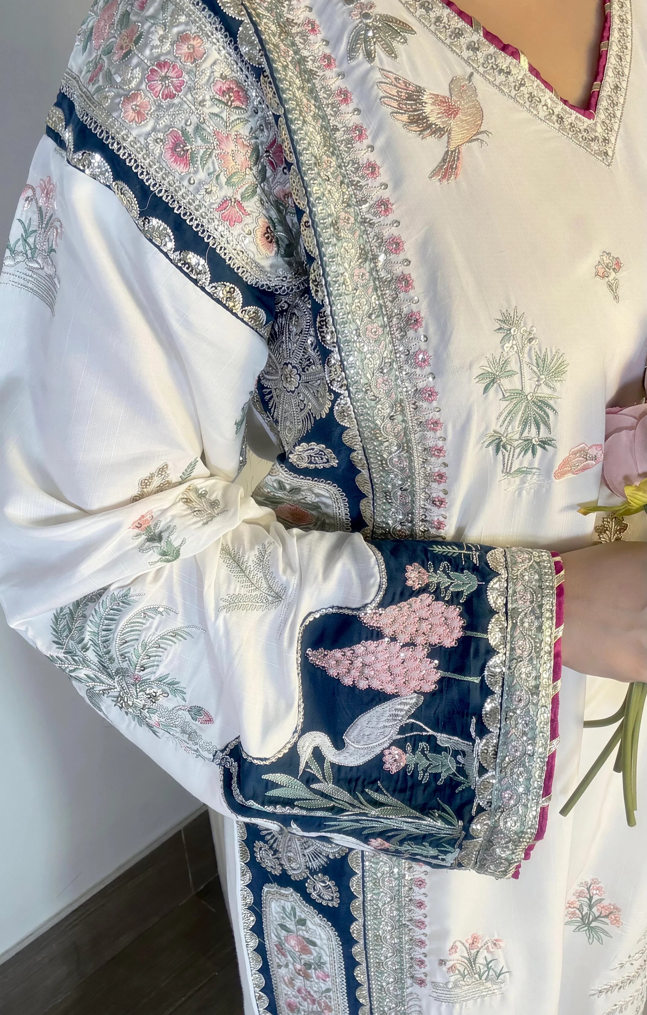 A close-up of a white traditional embroidered garment with navy blue borders, pink floral details, and bird motifs, worn by a person holding a pink flower.