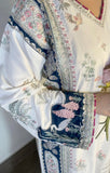 A close-up of a white traditional embroidered garment with navy blue borders, pink floral details, and bird motifs, worn by a person holding a pink flower.